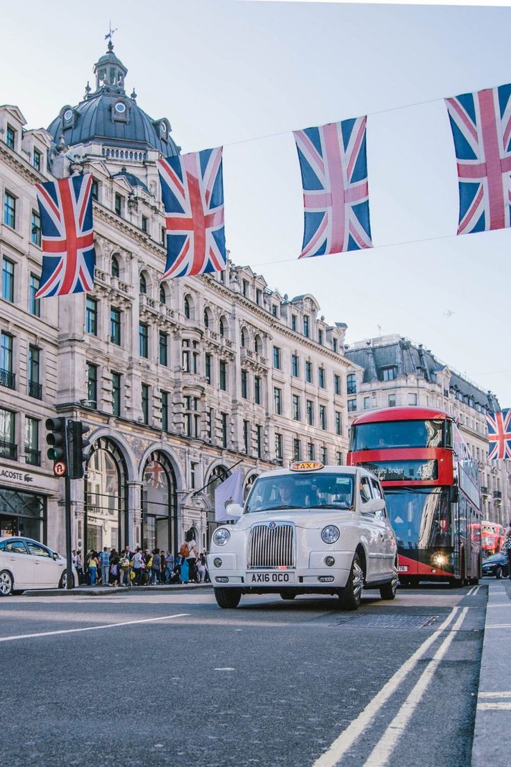 a double decker bus driving down a street next to tall buildings with flags hanging from it's sides