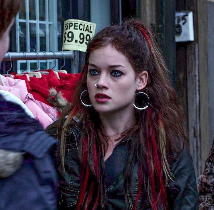 a woman with long red hair and piercings on her ear standing in front of a clothing rack