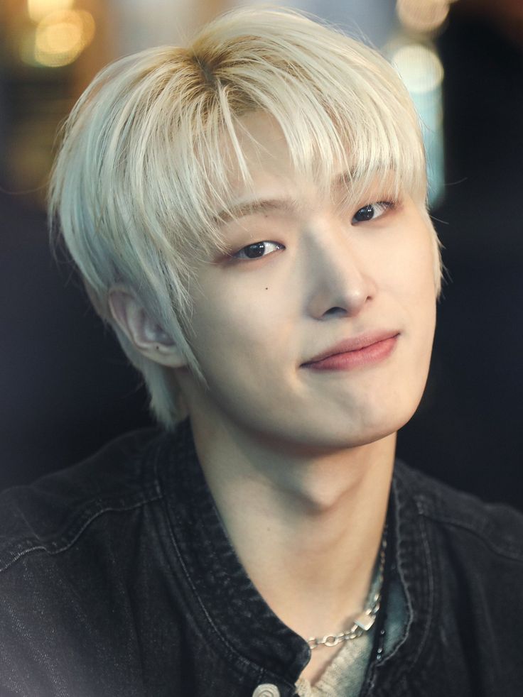 a young man with blonde hair wearing a black shirt and silver necklace looking at the camera