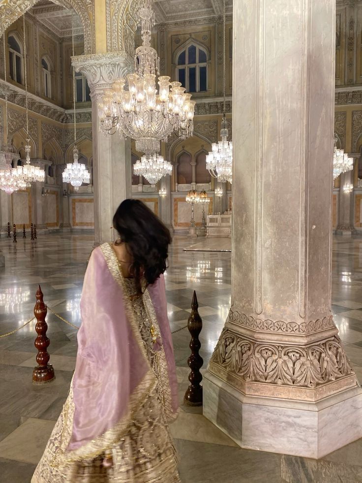 a woman in a pink and gold dress standing next to a pillar with chandeliers hanging from it