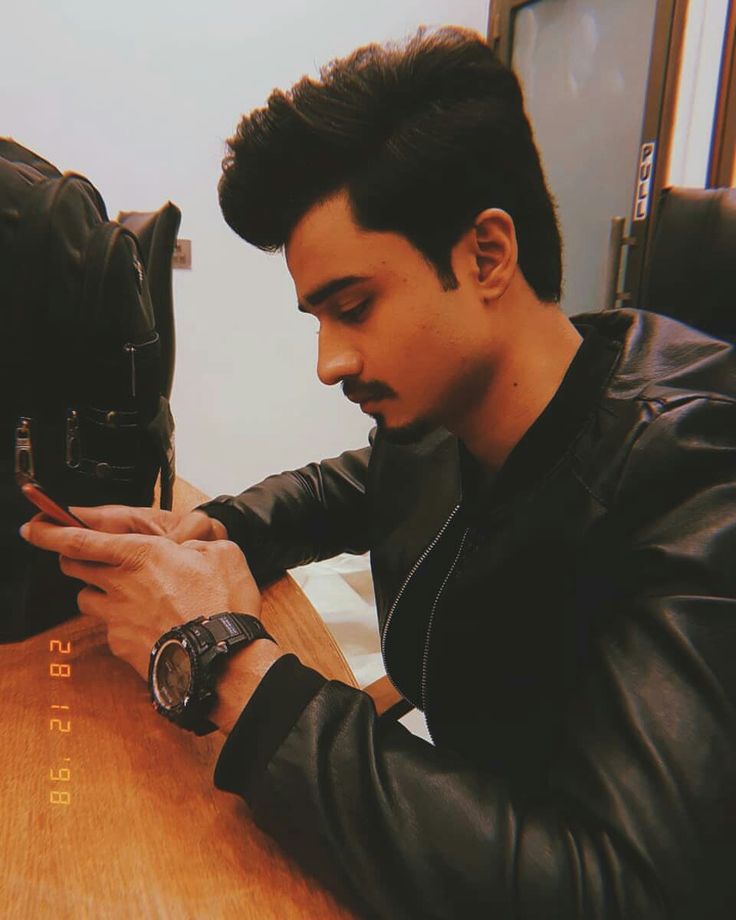 a young man sitting at a table looking at his cell phone while wearing a leather jacket