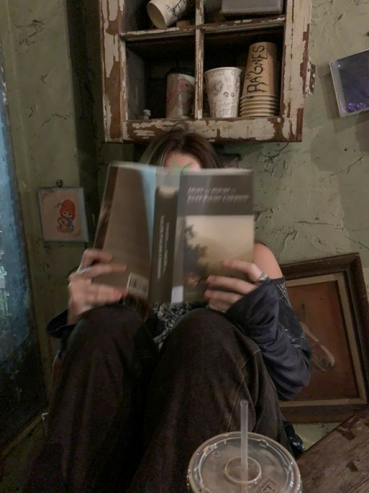 a woman is sitting on the floor reading a book and drinking from a glass in front of her