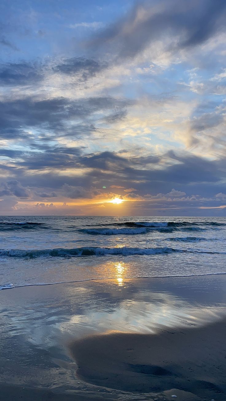 the sun is setting over the ocean with clouds reflected in the wet sand and water