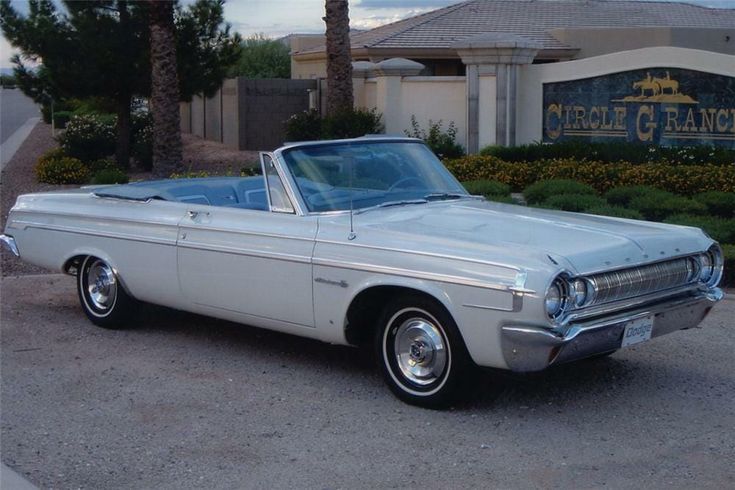 an old silver car parked in front of a hotel with palm trees and bushes behind it