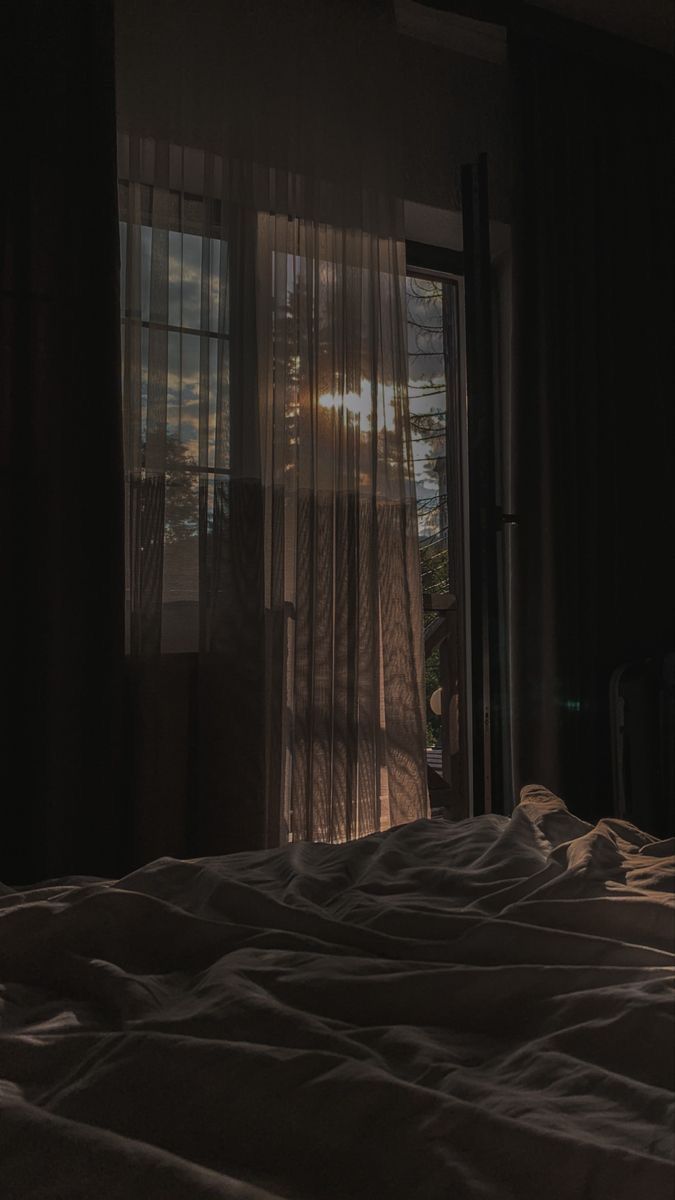 an unmade bed in front of a window with the sun shining through it's curtains