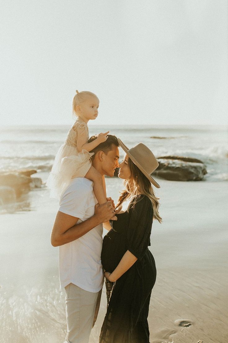 a woman holding a baby on her shoulders while standing next to a man in front of the ocean