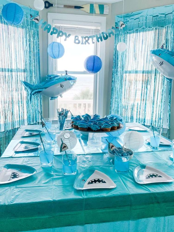 a birthday party table with blue decorations and paper fish on it's head, along with cupcakes