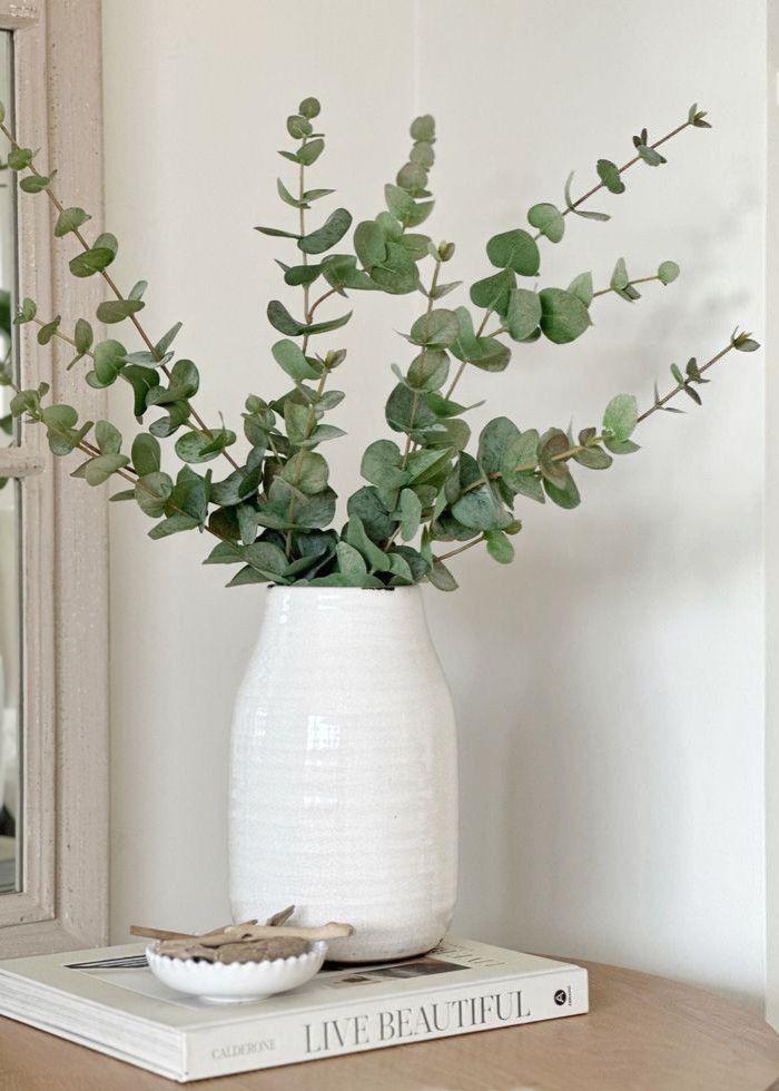 a white vase filled with green leaves on top of a wooden table next to a mirror