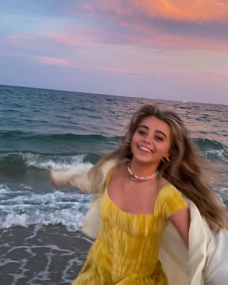 a woman in a yellow dress is walking on the beach with her arms out to the water