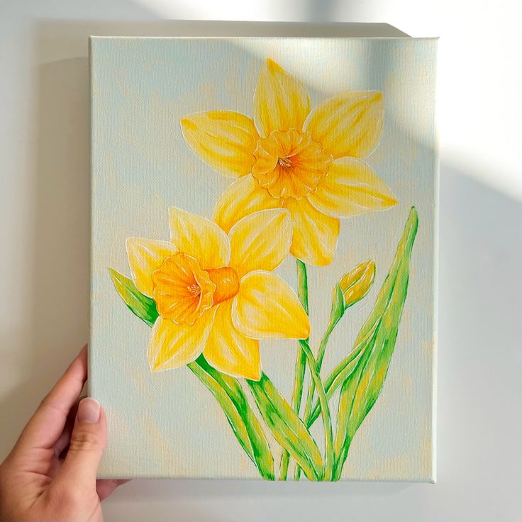 a painting of yellow flowers is being held up by a person's hand on a white surface