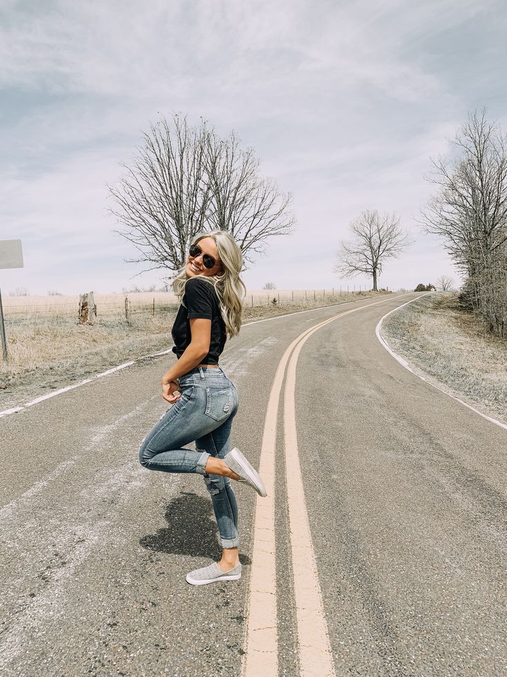 a woman in black shirt and jeans standing on the side of an empty road with trees