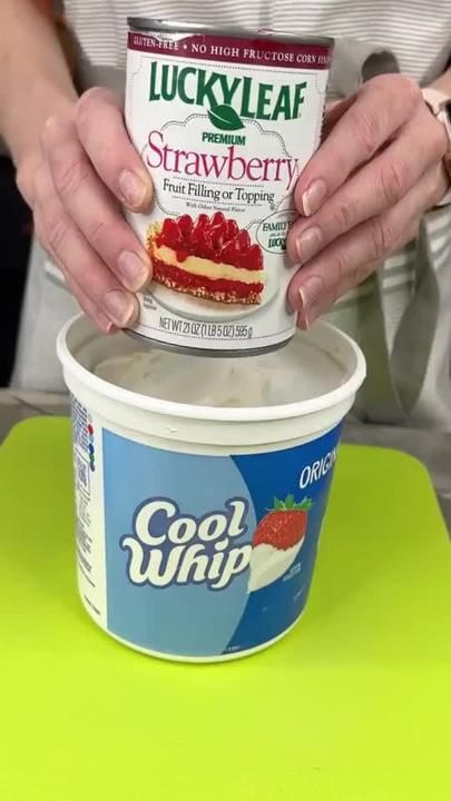 a person holding up a container of ice cream with strawberries on top and another cup in the background