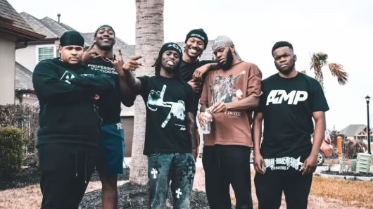 five young men standing together in front of a palm tree and some houses with their arms around each other