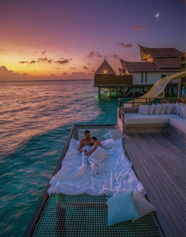 a man laying on top of a bed next to the ocean at sunset in front of an over water villa