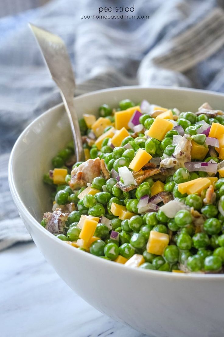 a white bowl filled with peas, cheese and other vegetables next to a silver spoon