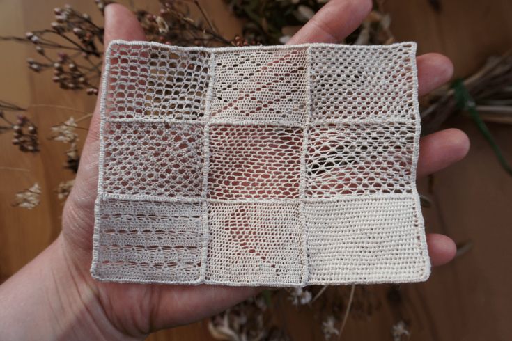 a hand holding a piece of white crocheted material with flowers in the background