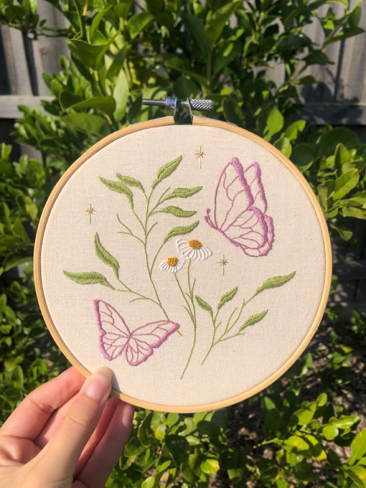 a hand holding up a embroidery hoop with some flowers and butterflies on it in front of bushes