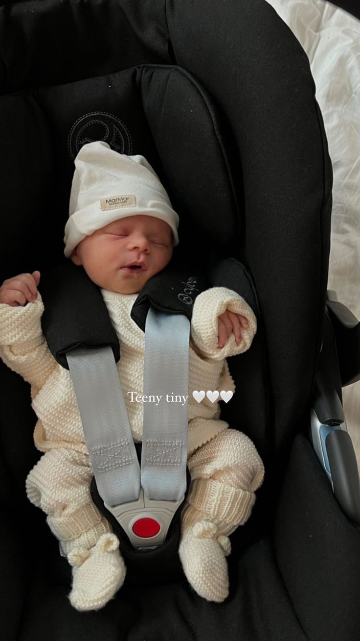 a baby in a car seat wearing a white hat and sweater with suspenders on