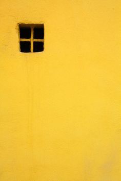 a yellow wall with a small window on the side and a black cat sitting in front of it