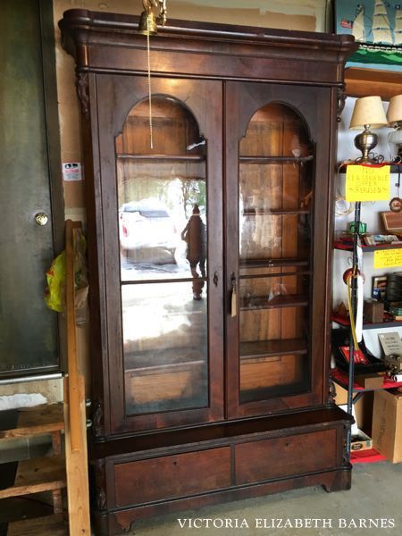 an old wooden china cabinet with glass doors in a room filled with shelves and other items