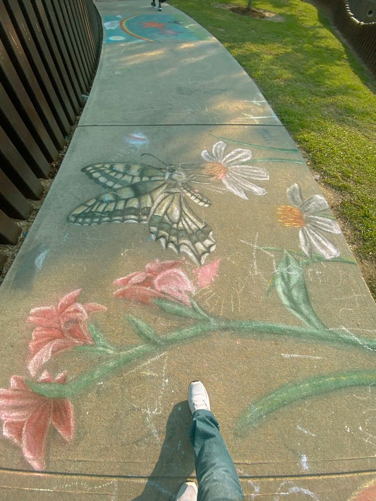 someone's feet are standing on the sidewalk painted with flowers and butterfly wings in front of a park bench