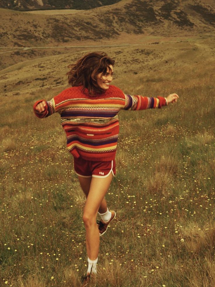 a woman is running in the grass with her arms spread out and she's wearing a colorful sweater