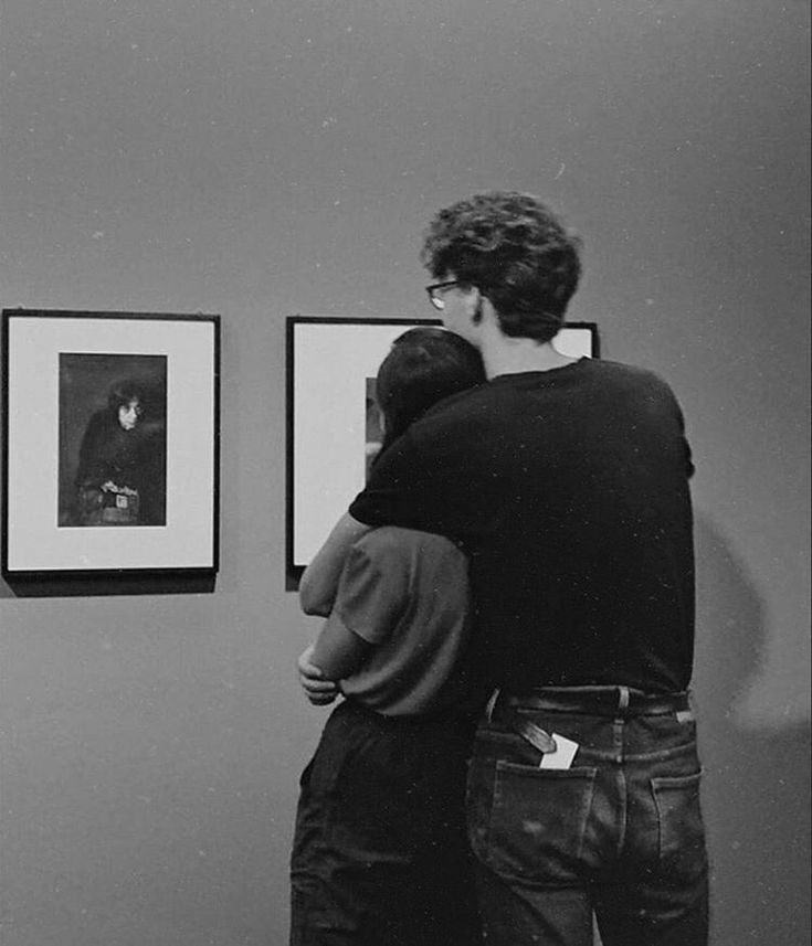 two people are looking at pictures on the wall in an art gallery, one is hugging the other's back