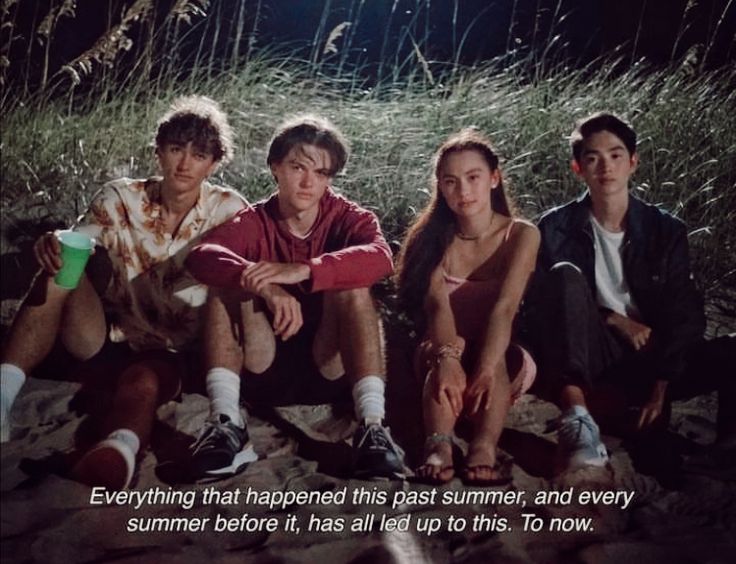 four people sitting on the ground in front of some tall grass and one person is looking at the camera