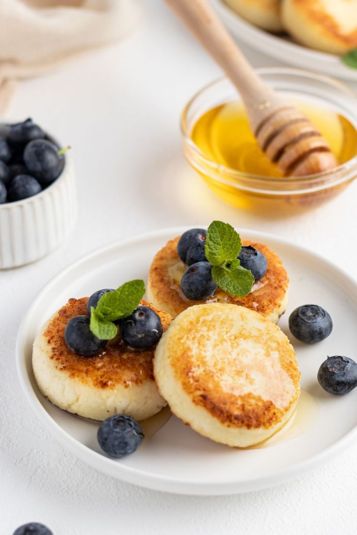 Syrniki served with honey, bluberries, and mint leaves on a white plate Ukrainian Breakfast Recipes, Syrniki Pancakes, Farmer Cheese Pancakes, Sirniki Russian, Syrnyky Recipe, Sirniki Recipe, Russian Food Recipes, Russian Food, Syrniki Recipe