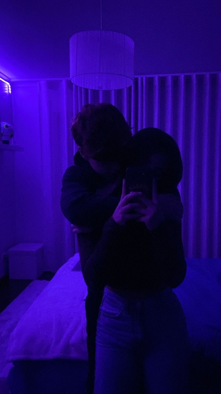 two people are hugging in the dark with purple lighting on them and one person is taking a photo