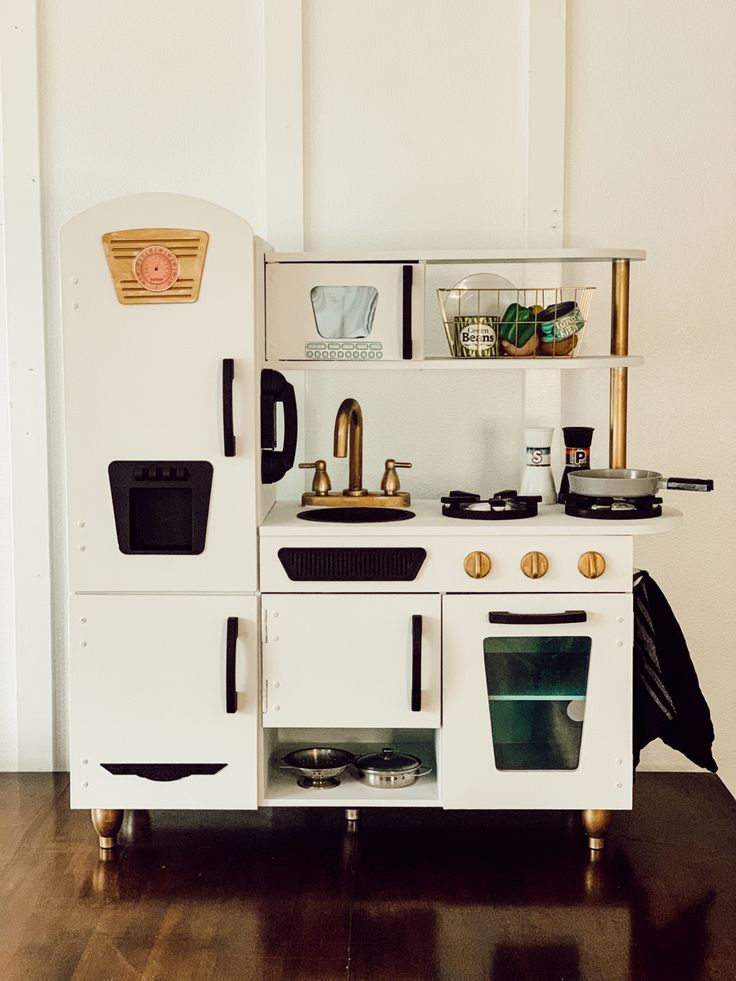 an old fashioned white wooden play kitchen with black and gold accessories on the top shelf