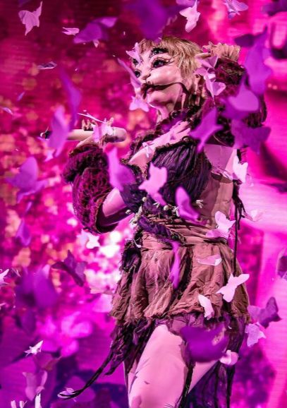 a woman dressed in costume standing on stage with purple butterflies around her and holding a microphone