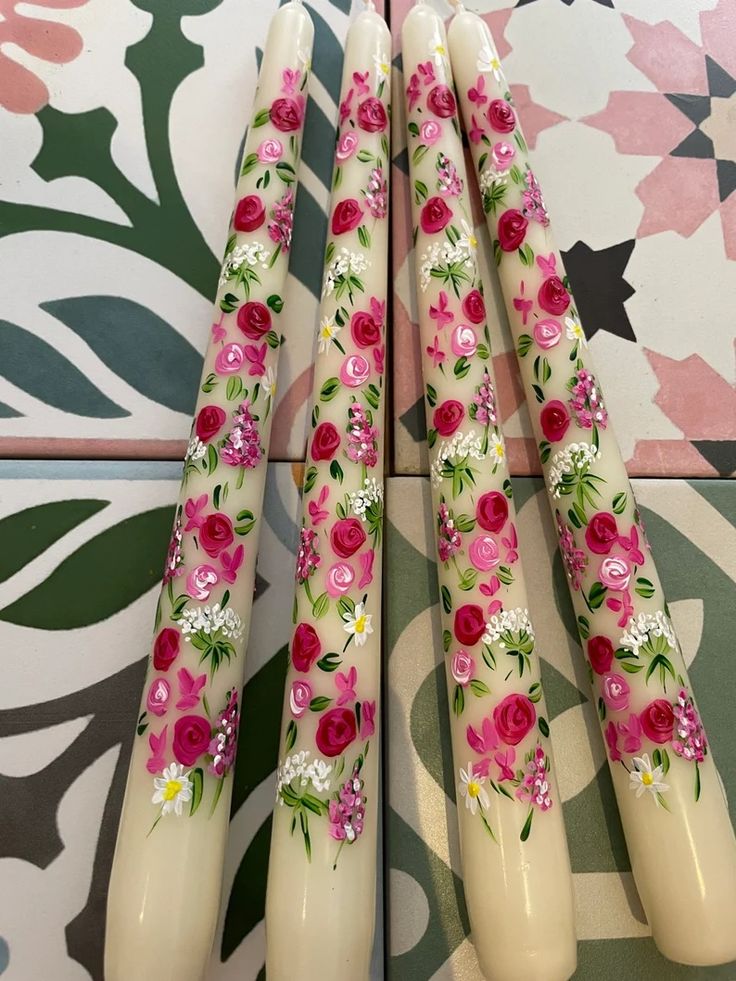 four candles with flowers painted on them sitting on a patterned tablecloth, one is white and the other is pink