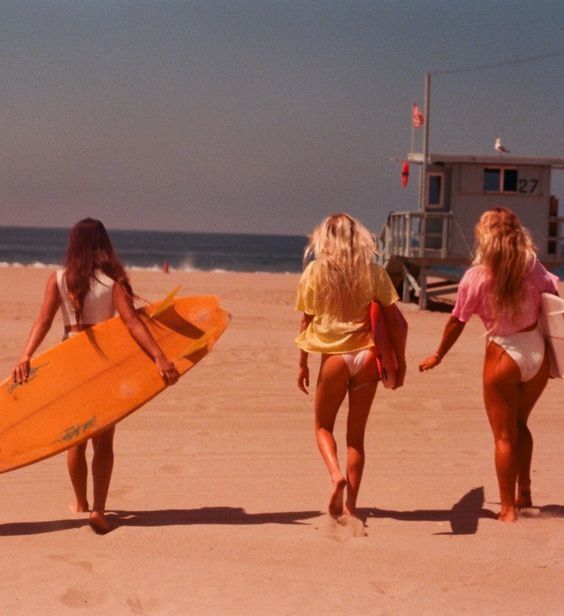 three women walking on the beach with their surfboards in hand and one holding a yellow board