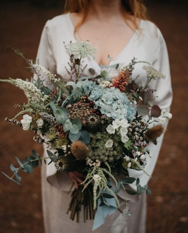 a woman holding a bouquet of flowers and greenery on her wedding day in the woods