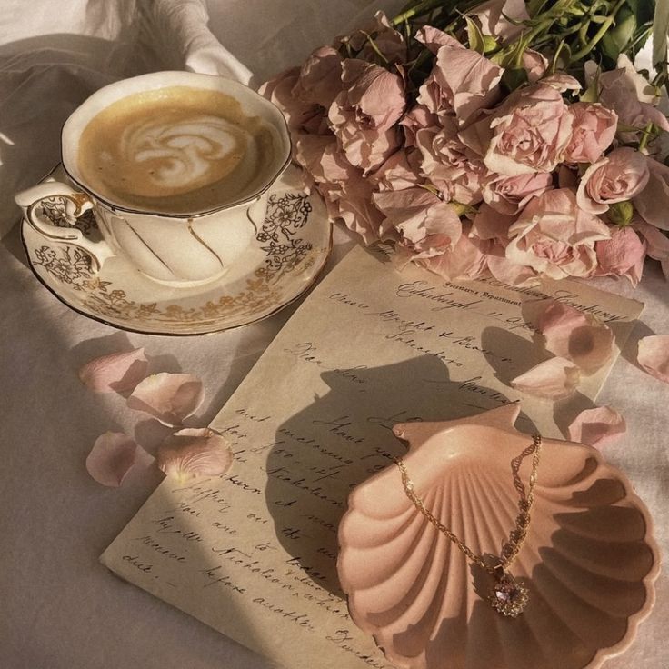 a cup of coffee next to a seashell and pink flowers on a white table cloth