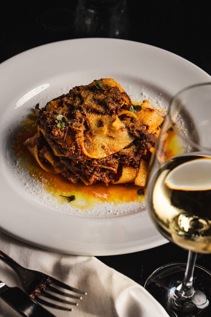 a white plate topped with lasagna next to a glass of wine and silverware