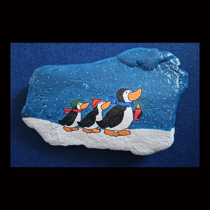 painted rock with penguins on it sitting on a blue surface in front of a black background