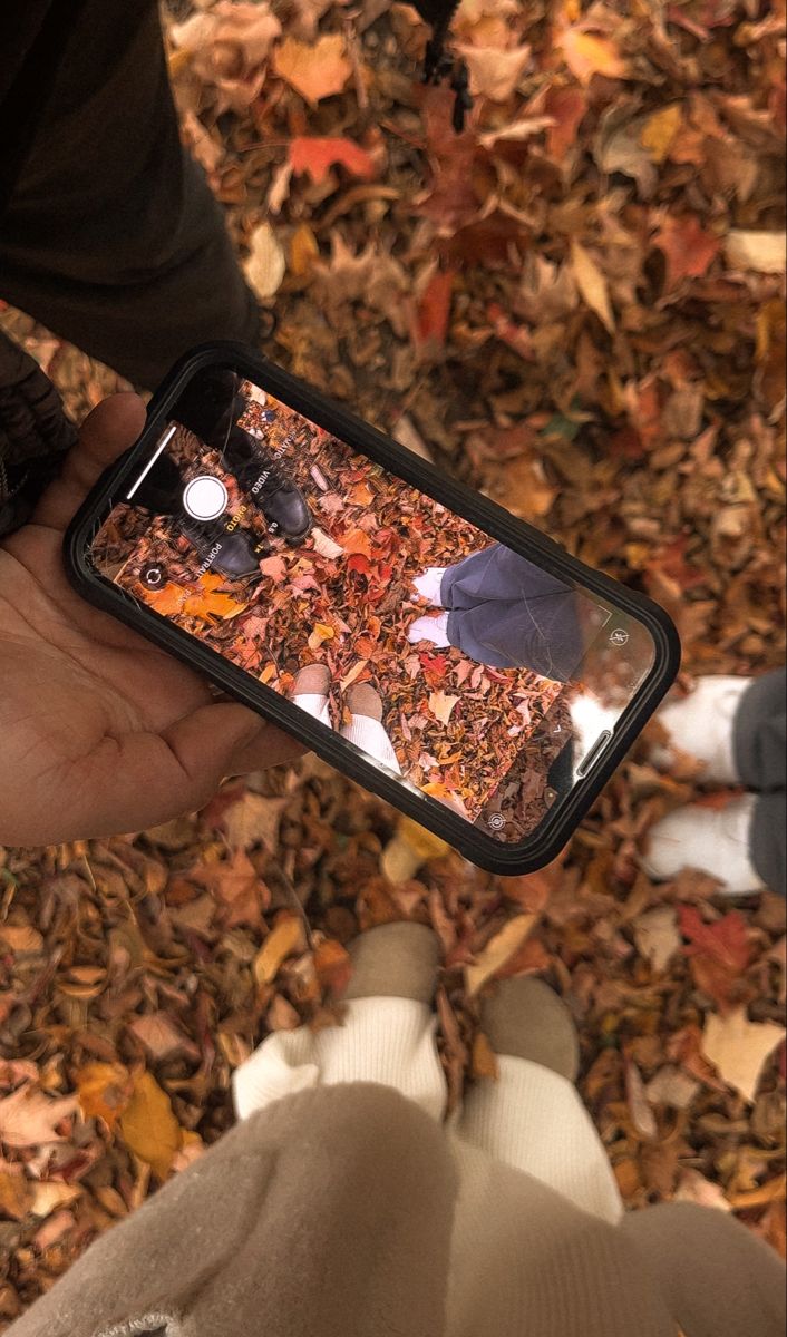 someone is holding their cell phone in front of the ground with leaves on it,