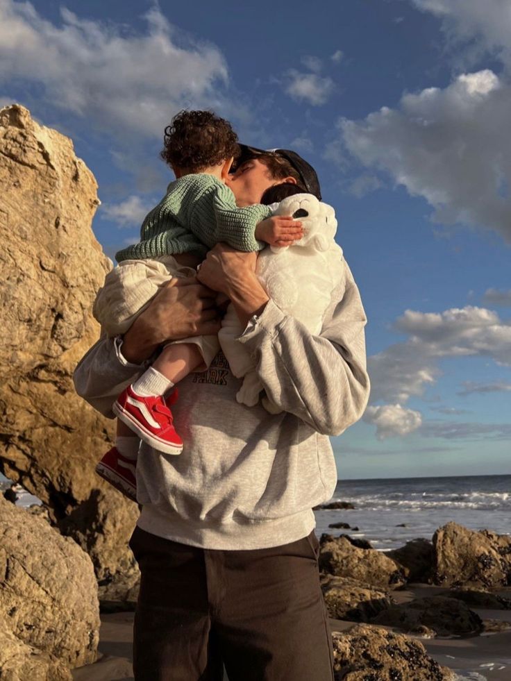 a man holding a child on the beach with rocks in the background and clouds in the sky