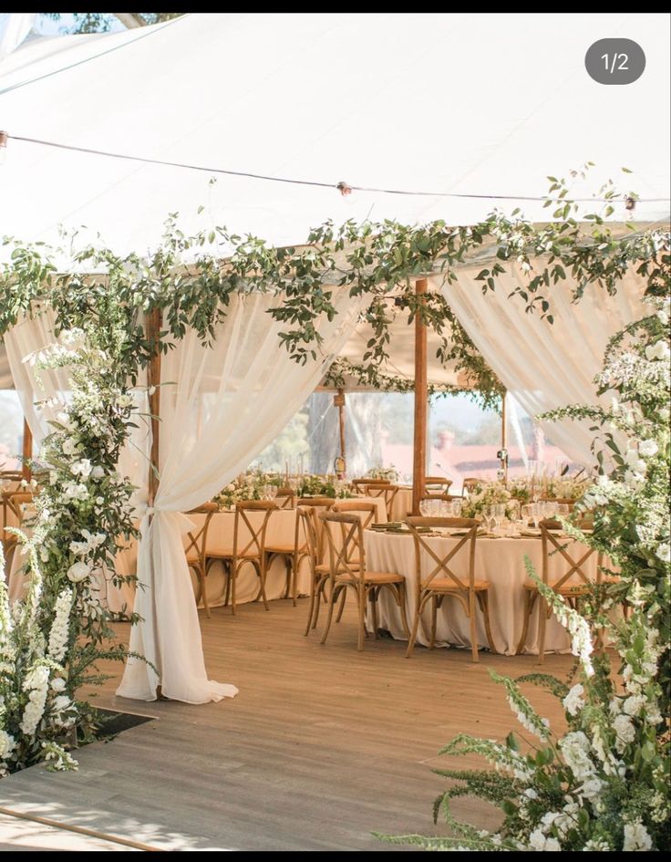 the inside of a tent with tables and chairs covered in white drapes, flowers and greenery