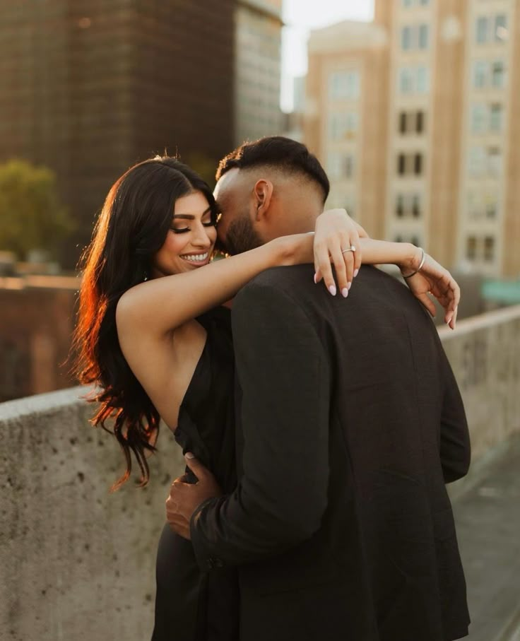 a man and woman embracing each other while standing on a bridge in front of tall buildings