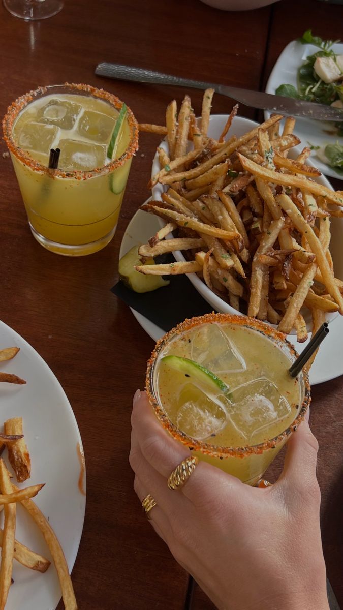 two people are holding glasses with drinks and french fries on the table in front of them