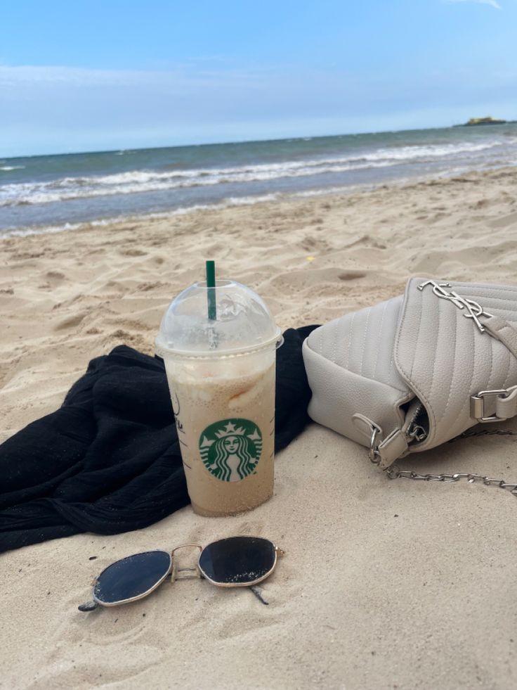 a starbucks drink sitting on top of a sandy beach next to sunglasses and a purse