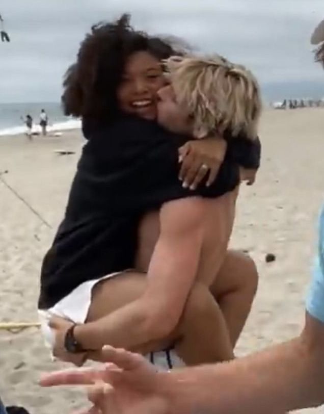 two women are hugging on the beach while one woman is pointing at something in her hand