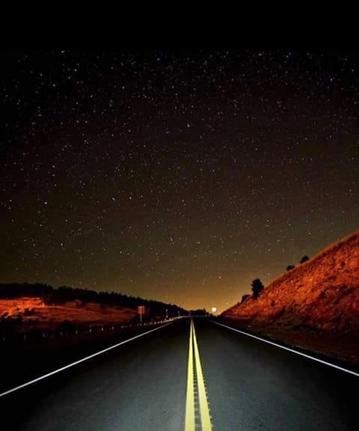 an empty road at night with the stars in the sky and some hills behind it