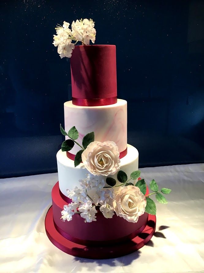 a three tiered cake with white flowers and leaves on the top, sitting on a red stand