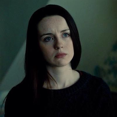 a woman with dark hair and blue eyes stares at the camera while standing in a dimly lit room