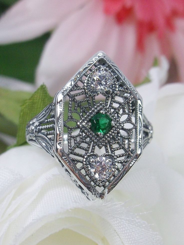 Simulated Green Emerald & White Cubic Zirconia (CZ) Sterling Silver RingCharlotte Design#D231 Resurrected from the Art Deco movement, this gorgeous Art Deco reproduction filigree ring is crafted in sterling silver. This lovely filigree ring is set with a stunning quality 2mm center round-cut simulated green emerald gemstone. Two small white cubic zirconia gemstones also grace the face of the ring, one near each point, each set in the center of a tiny heart. There are 2 filigree accent flowers th Art Deco Silver Emerald Wedding Ring, Silver Art Deco Emerald Wedding Ring, Silver Emerald Ring With Intricate Design For Anniversary, Ornate Silver Diamond Ring With Accents, Vintage Silver Filigree Ring With Diamond Accents, Ornate Silver Diamond-cut Rings, Silver Rings With Intricate Cubic Zirconia Design, Anniversary Silver Emerald Ring With Intricate Design, Heirloom Sterling Silver Filigree Ring With Diamond Accents