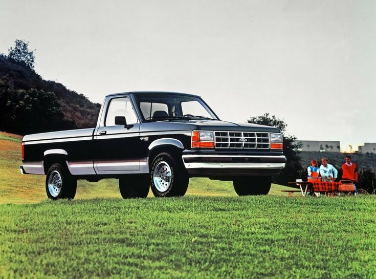 an old black pickup truck parked in a field with people sitting on the grass behind it